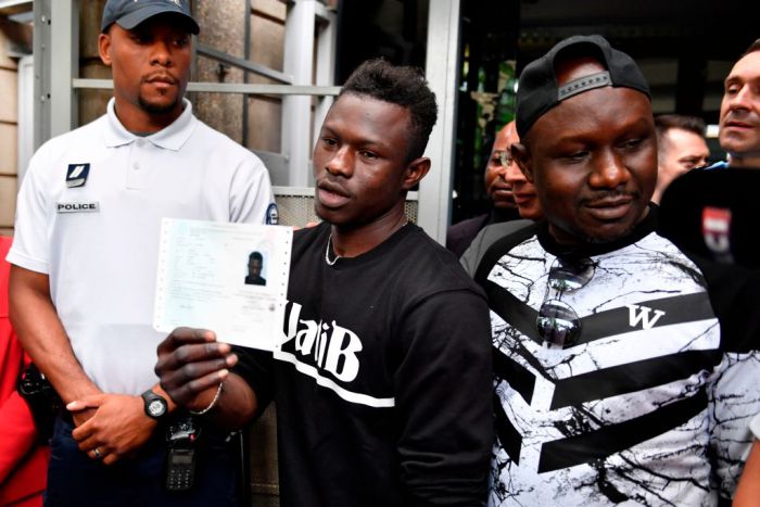 Malian migrant Mamoudou Gassama (C) flanked by his older brother (R) holds his temporary residence permit after receiving it at the Prefecture of Bobigny, northeast of Paris on May 29, 2018, one day after he was honored by the French President for scaling an apartment building to save a 4-year-old child dangling from a fourth-floor balcony. - Two days after his daring rescue -- viewed millions of times online -- Mamoudou Gassama, 22-year old,  nicknamed "Spiderman" by French media for his astonishing climbing ability and feted as a hero, was offered by French President citizenship, and a job with the fire service. 